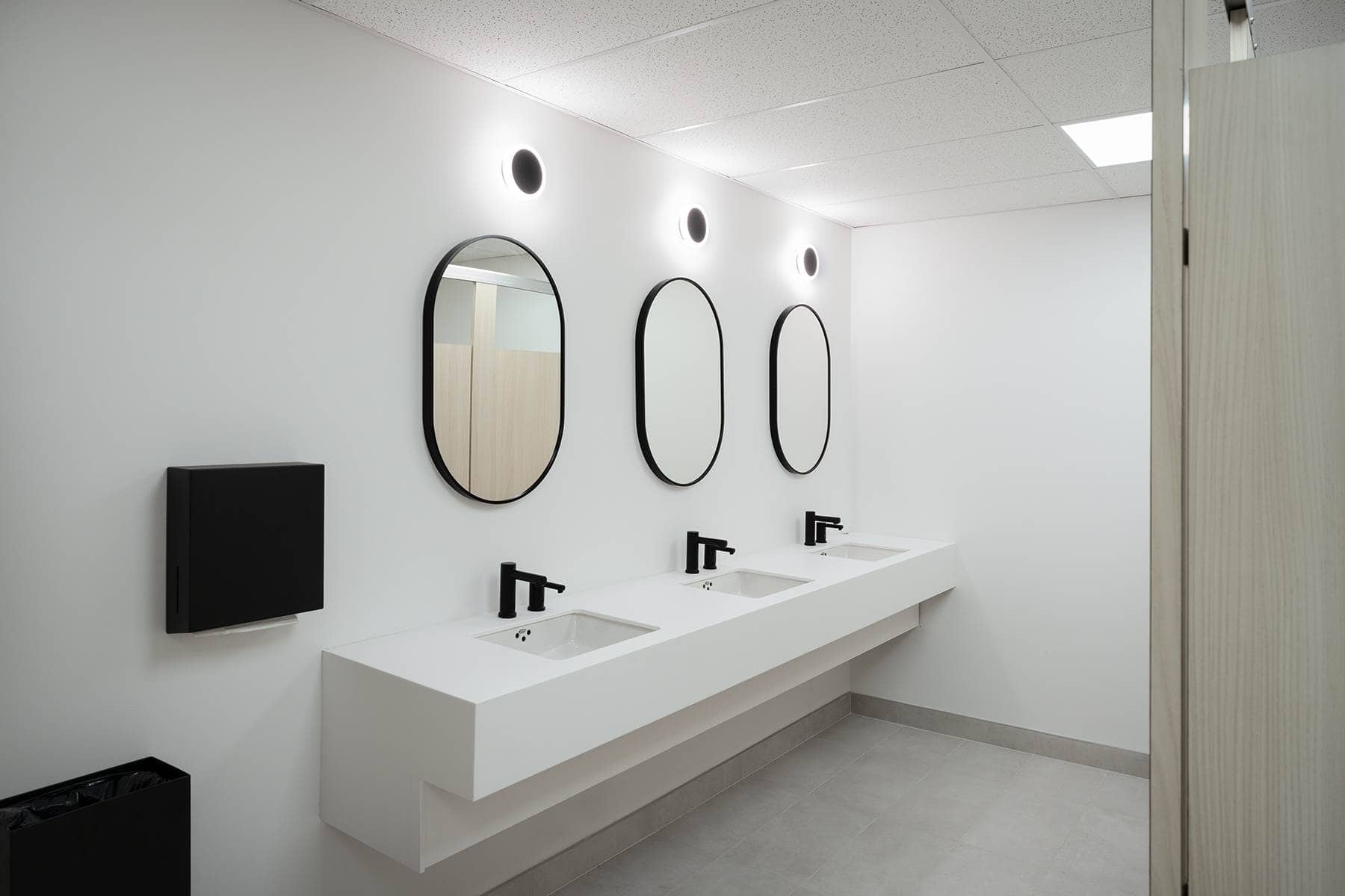 Des toilettes publiques modernes avec un comptoir d'évier blanc, trois miroirs ovales et des robinets noirs, dotés d'un éclairage zénithal lumineux.