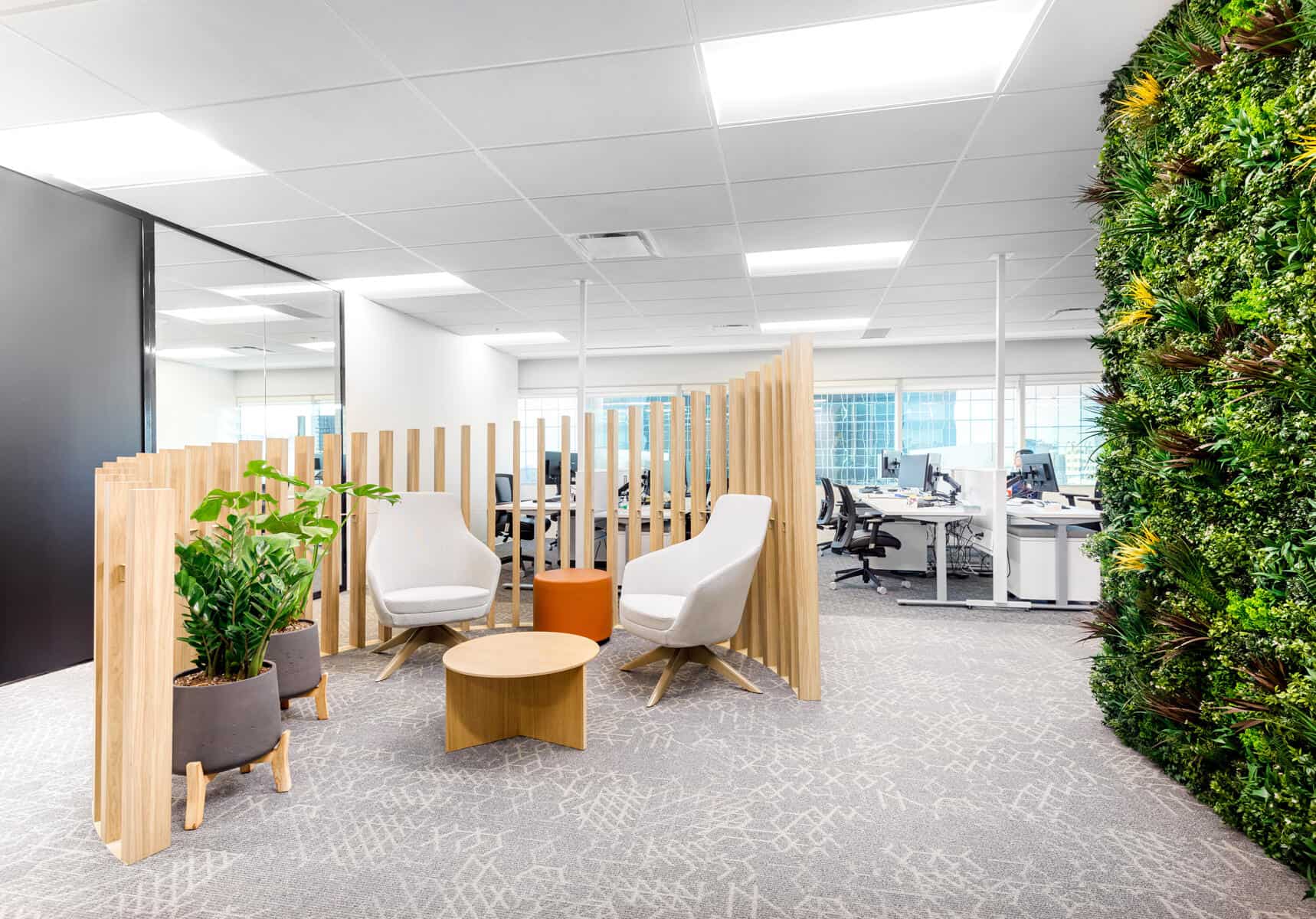 Rénovation de bureau avec un intérieur moderne avec un coin salon comprenant des chaises blanches, des cloisons en bois, un mur végétal vert et des bureaux avec des ordinateurs en arrière-plan.