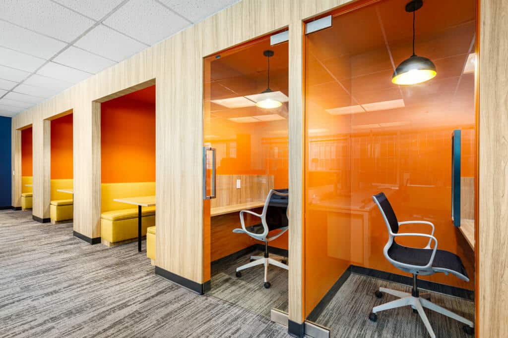 Une rangée de cabines avec des portes vitrées encastrées dans un mur, une avec des bancs pour les réunions et deux avec des chaises de travail et des bureaux