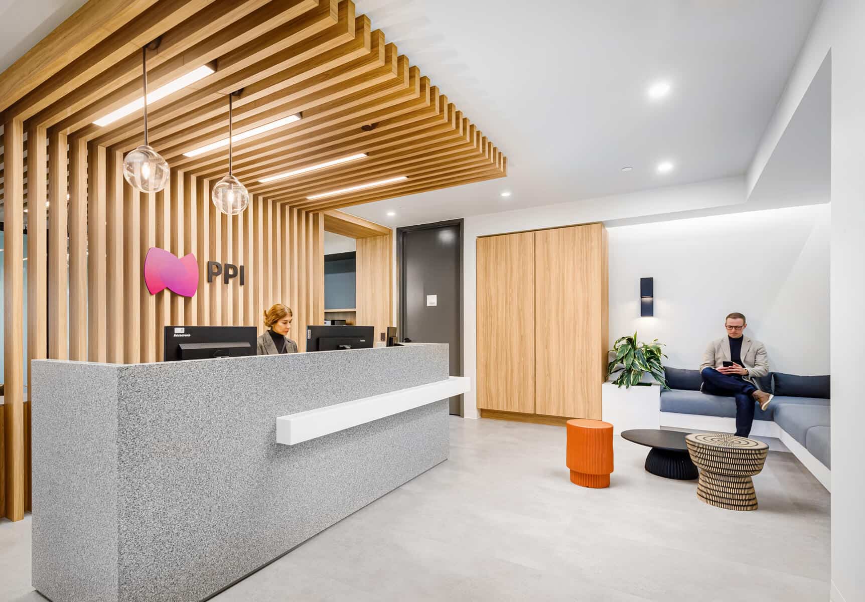 Espace de réception de bureau moderne avec un homme et une femme, des accents en bois élégants, un bureau d'entrée, un coin salon et des suspensions suite à une rénovation de bureau.
