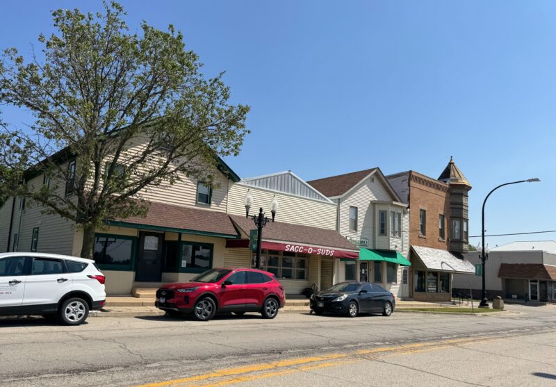 4 bâtiments d'affilée et trois semblent vides, dans une rue du centre-ville de Waterman, dans l'Illinois. Le bâtiment complet est la laverie Sacc O Suds avec plusieurs voitures garées devant. Le bâtiment au bout de la brique avec une tourelle et un logement au deuxième étage. Les trois autres sont en structure de 2 étages, avec un espace pour le commerce de détail en bas et des baies vitrées.