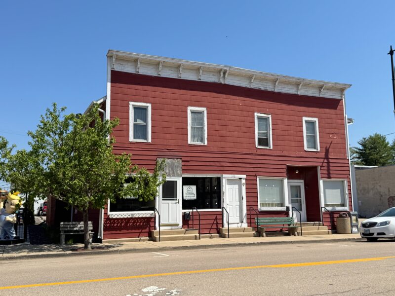 Immeuble de deux étages du centre-ville de Hinckley, dans l'Illinois, avec un revêtement de couleur bordeaux. Trois portes d'entrée avec 2 marches devant chacune et 4 baies vitrées. 4 petites fenêtres à l'étage avec rideaux blancs anciens. Il y a un banc à gauche, un au milieu et une poubelle à droite. Le bâtiment semble être une structure de la fin des années 1800 ou du début des années 1900. Les trois devantures de magasins semblent vides.