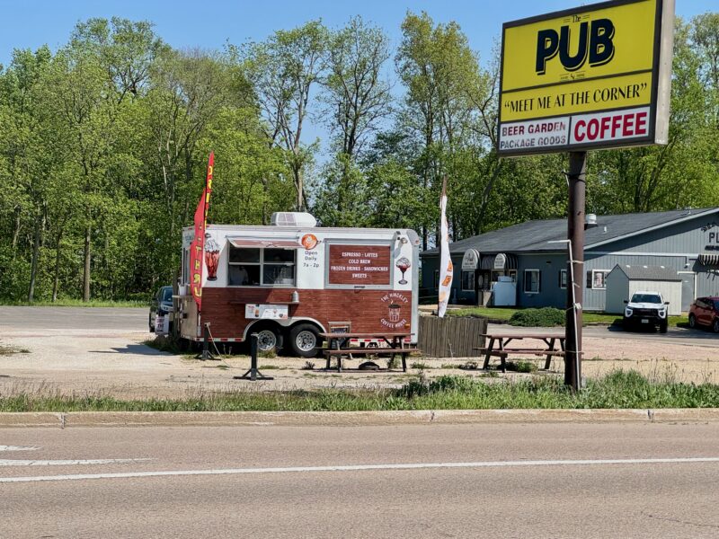 Roulotte marron et blanche transformée en café dans un parking. Deux bancs de pique-nique devant et deux panneaux en tissu fluide sur des poteaux. L’herbe le long des bordures doit être coupée. Il y a un panneau le long de la route avec The Pub en noir sur fond jaune et "rendez-vous au coin" en dessous de ça. Au bas du panneau se trouvent « beer garden » et « café » en lettres rouges sur fond blanc. au fond du parking, sur la droite, se trouve une structure grise d'un étage qui semble être le pub.