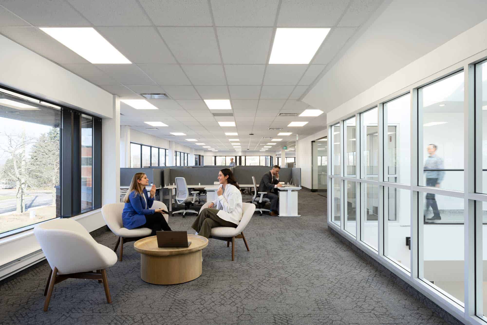 Conception de bureau moderne avec des employ&eacute;s travaillant &agrave; des bureaux et deux femmes assises, discutant dans un coin salon pr&egrave;s de grandes fen&ecirc;tres.