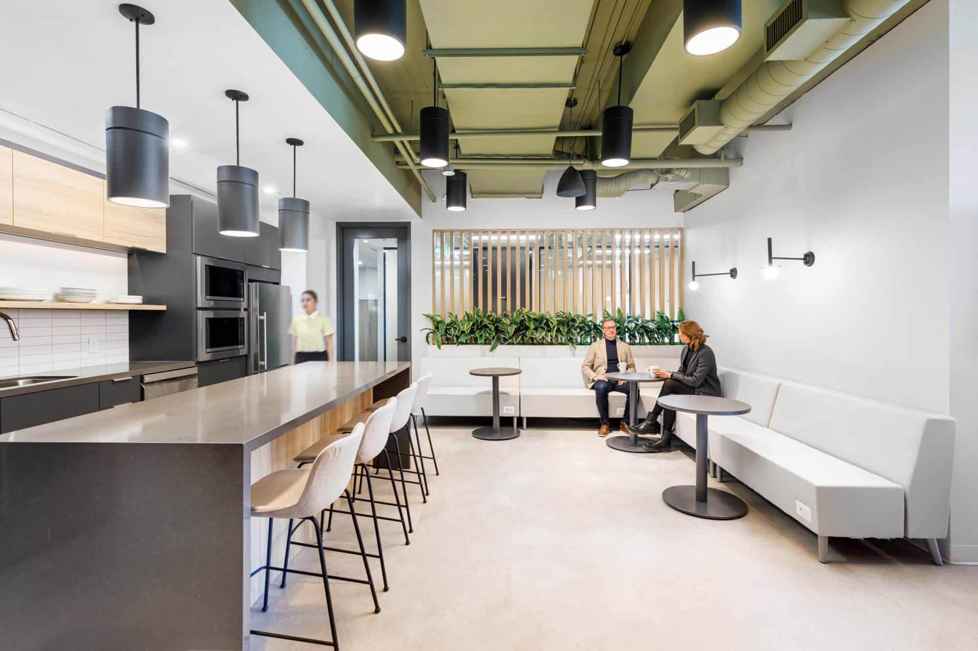 Conception et construction de bureaux modernes comprenant une cuisine et un coin salon avec des personnes assises et parlant, pr&eacute;sentant une d&eacute;coration int&eacute;rieure &eacute;l&eacute;gante &eacute;clair&eacute;e par la lumi&egrave;re naturelle, des accents verts et des tables communes.
