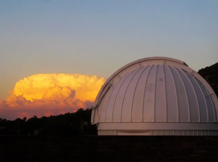 Un dôme d'observatoire blanc sur un fond montagneux avec des nuages orageux ensoleillés à l'horizon.